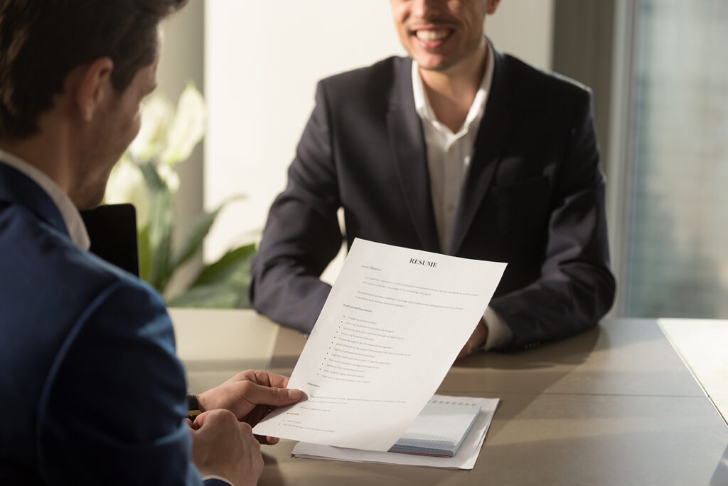 Friendly employer conducting job interview, reviewing good resume of prepared skilled smiling applicant waiting for result at background, recruiter considering cv, focus on document, close up view