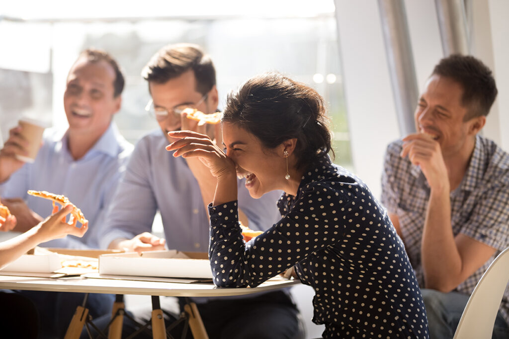 Woman laughing at funny joke eating pizza with diverse coworkers in office, friendly work team enjoying positive emotions and lunch together, happy colleagues staff group having fun at break