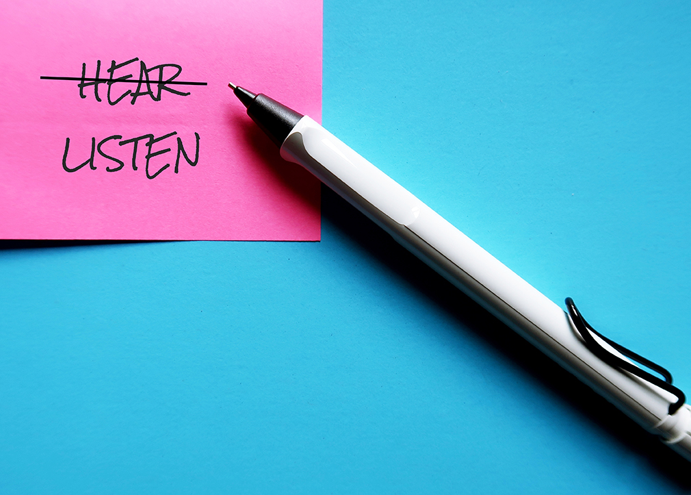 Pen and pink note on blue background with handwritten text HEAR changed to LISTEN, means to be better listener with empathy, not just hearing, to see the world in speaker eyes and understand more