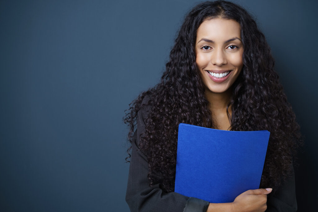 Motivated young African American job seeker clutching a blue CV in her hands standing against a blue background with copy space beaming at the camera