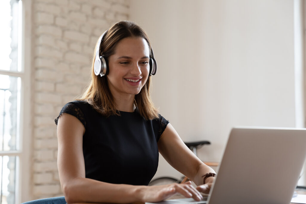 Smiling woman wearing wireless headphones working typing on notebook sit at desk in office workplace. Enjoy e-learning process, easy comfortable application usage, listen music during workday concept