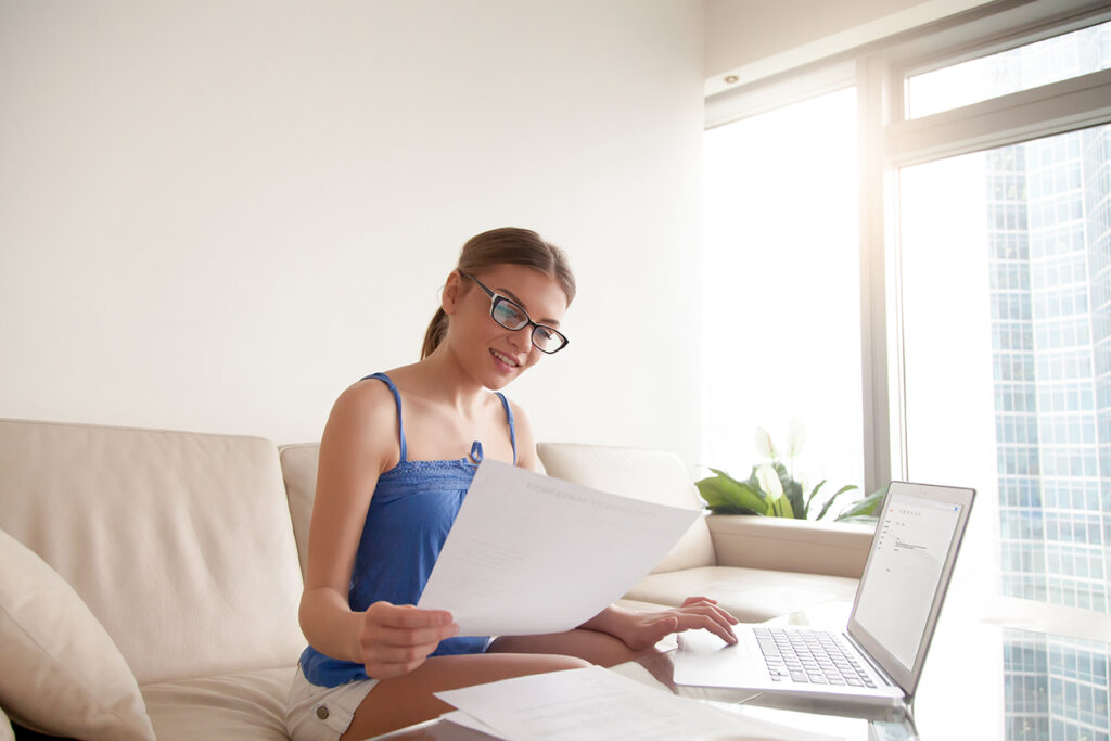 Young woman wearing glasses holding business document using laptop, hr manager working at home composing e-mail job offer to new open vacancy candidate, typing answer letter after resume consideration